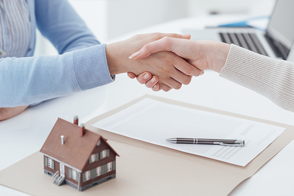 An image of two women shaking hands with a miniature house in the foreground.